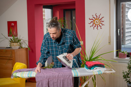 man with iron on ironing board with clothers in living roomの写真素材