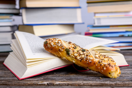 book and little whole grain bread in front of piles of different books on wooden tableの写真素材