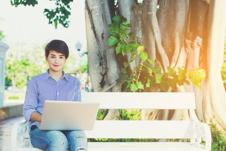 Beautiful short hair woman sitting on white chair under the tree and using laptop for outdoor working working her business in the park.のeditorial素材