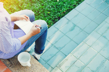 Body of woman sitting on the floor and writing a note in notebook  with favorite beverage in white cup beside between her outdoor working business in the park.のeditorial素材