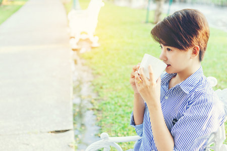Beautiful short hair woman sitting on a white chair and drinking a favorite beverage in white cup for resting between her outdoor working business in the park.の写真素材