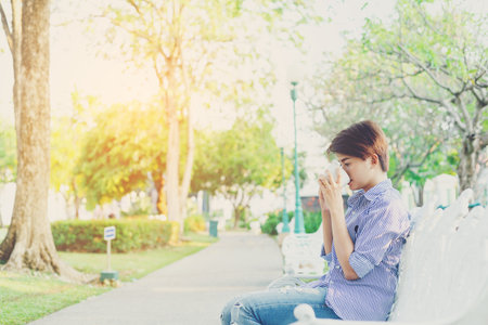 Beautiful short hair woman sitting on a white chair and drinking a favorite beverage in white cup for resting between her outdoor working business in the park.の写真素材