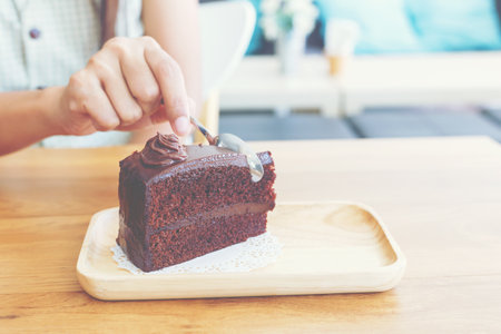 Hand of woman using a teaspoon for cut a piece of slice chocolate cake on wooden dish on the table of the cafe.の写真素材