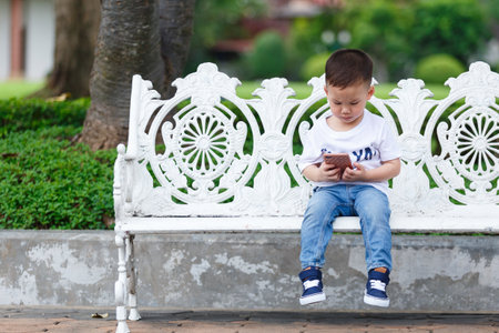 Little boy concentrate at smartphone and sit at white chair at the park.の写真素材