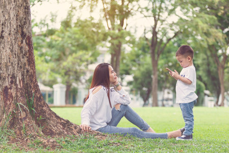 Little boy concentrate at smartphone with his mom sit together on green yard under giant tree.の写真素材