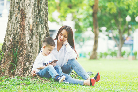 Little boy concentrate at smartphone with his mom sit together on green yard under giant tree.の写真素材
