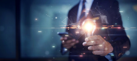 Businessman holding a bright glowing light bulb It is an inspiration for developing new creative ideas or imaginations. Polygons connect various brainstorming ideas on a dark blue background.の写真素材