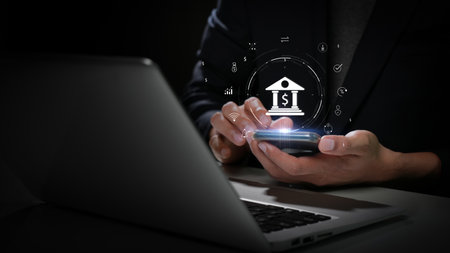 Businessman using smartphone for mobile banking with digital financial icons, sitting in front of a laptop on dark background.の写真素材
