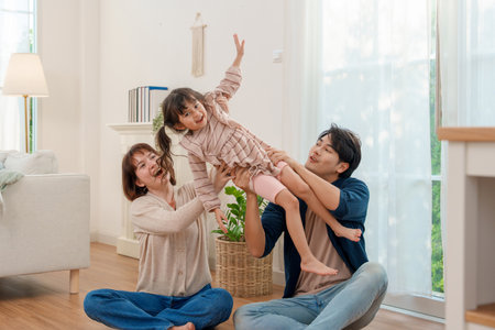 Joyful Asian parents sitting on floor, lifting daughter high in bright living room. Family sharing playful, heartwarming moment of love and fun.の写真素材