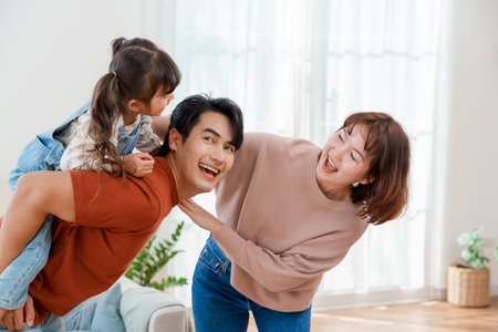 Happy Asian family having fun at home with father giving a piggyback ride to his little daughter while the mother smiles with joy Lifestyle family bonding and togetherness conceptの写真素材