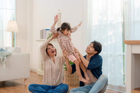 Asian parents sitting on floor, lifting excited daughter high in bright living room. Family sharing playful, heartwarming moment of love and fun together.の写真素材