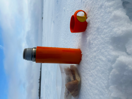 An orange flask and a plastic cup with a hot drink stands on the snow in winter.の写真素材