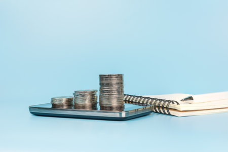 Coins stack on smartphone with notebook and pen on light blue background. business and financial planning concept.の写真素材