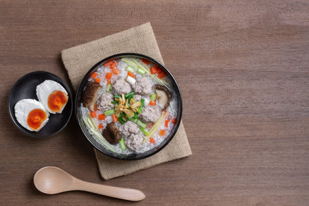 top view of boiled rice clear soup with pork in a ceramic bowl on wooden table, served with salted duck egg. homemade style food concept.の写真素材