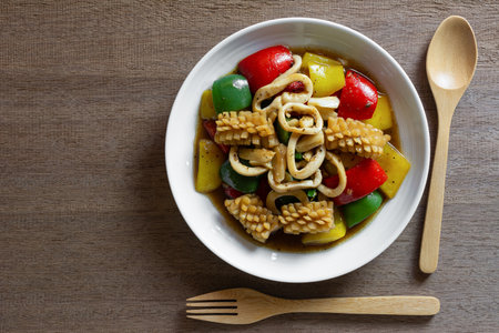 top view of stir fried squid with black and sweet pepper in a ceramic dish on wooden table. homemade...