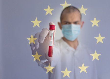 Coronavirus conceptual photo with doctor wearing a medical face mask and gloves, holding a blood test result for the COVID-19 pandemic and European Union flag, blurred backgroundの写真素材