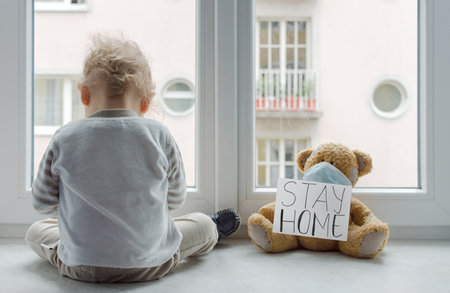 Child in home quarantine playing at the window with his sick teddy bear wearing a medical mask against viruses during coronavirus and flu outbreak, with the advice to stay home.の写真素材