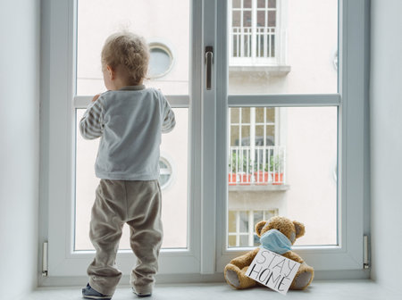 Child in home quarantine playing at the window with his sick teddy bear wearing a medical mask against viruses during coronavirus and flu outbreak, with the advice to stay home.の写真素材