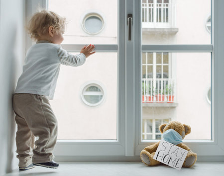 Child in home quarantine playing at the window with his sick teddy bear wearing a medical mask against viruses during coronavirus and flu outbreak, with the advice to stay home.の写真素材