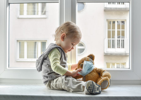Child in home quarantine playing at the window with his sick teddy bear wearing a medical mask against viruses during coronavirus and flu outbreak. Children and illness COVID-2019 disease conceptの写真素材