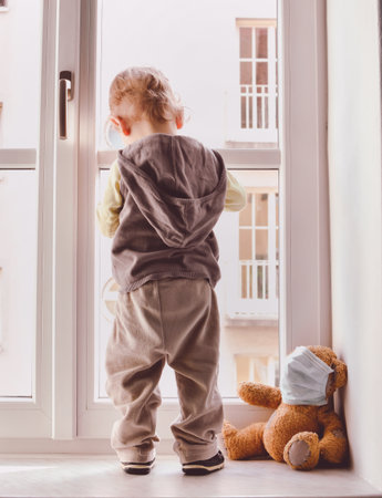 Child in home quarantine looking out of the window with his sick teddy bear wearing a medical mask against viruses during coronavirus and flu outbreak. Children and illness COVID-2019 disease conceptの写真素材