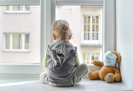 Child in home quarantine looking out of the window with his sick teddy bear wearing a medical mask against viruses during coronavirus and flu outbreak. Children and illness COVID-2019 disease conceptの写真素材