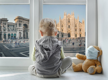 Milan/Italy - March 05, 2020: Child in home quarantine looking out of the window with his sick teddy bear at Milan Cathedral during coronavirus COVID-19 and flu outbreak.の写真素材