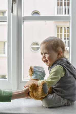 Child in home quarantine playing at the window with his sick teddy bear wearing a medical mask against viruses during coronavirus COVID-2019 and flu outbreak. Children and illness disease concept.の写真素材