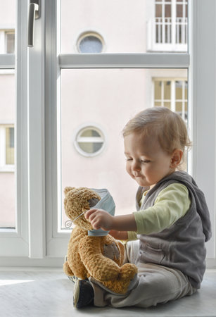 Child in home quarantine playing at the window with his sick teddy bear wearing a medical mask against viruses during coronavirus COVID-2019 and flu outbreak. Children and illness disease concept.の写真素材
