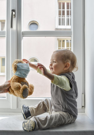 Child in home quarantine playing at the window with his sick teddy bear wearing a medical mask against viruses during coronavirus COVID-2019 and flu outbreak. Children and illness disease concept.の写真素材