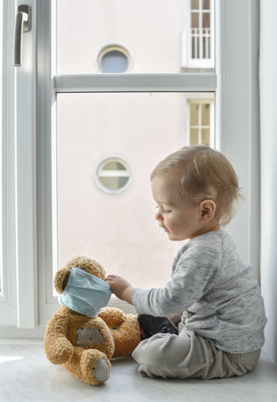 Child in home quarantine playing at the window with his sick teddy bear wearing a medical mask...