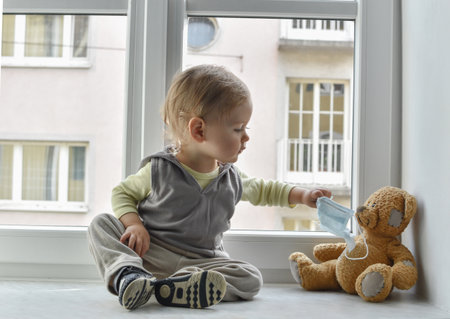 Child in home quarantine at the window putting a medical mask on his sick teddy bear, for protection against viruses during coronavirus COVID-19 and flu outbreak. Children and illness disease conceptの写真素材