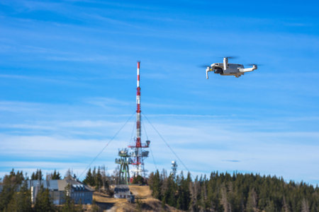 White drone with photo camera flying in the blue sky and telecommunication tower in the backgroundの写真素材