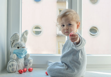 Easter 2020 concept with child in home quarantine playing at the window with his sick plush bunny wearing a medical mask against viruses during coronavirus COVID-2019 and flu outbreak.の写真素材
