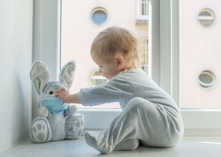 Easter 2020 concept with child in home quarantine playing at the window with his sick plush bunny wearing a medical mask against viruses during coronavirus COVID-2019 and flu outbreak.の写真素材