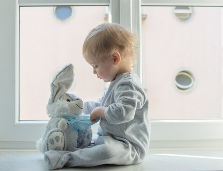 Easter 2020 concept with child in home quarantine playing at the window with his sick plush bunny wearing a medical mask against viruses during coronavirus COVID-2019 and flu outbreak.の写真素材