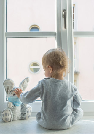Easter 2020 concept with child in home quarantine playing at the window with his sick plush bunny wearing a medical mask against viruses during coronavirus COVID-2019 and flu outbreak.の写真素材