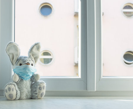 Easter 2020 concept with a plush bunny in home quarantine, at the window, wearing a medical mask against viruses during coronavirus COVID-2019 and flu outbreak.の写真素材