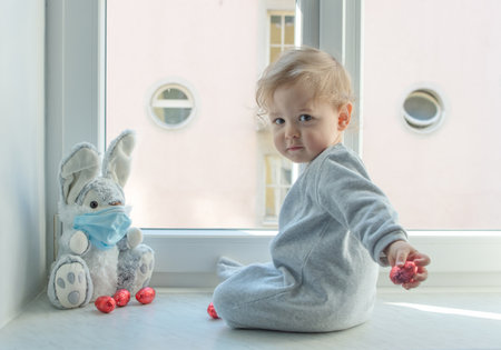 Easter 2020 concept with child in home quarantine playing at the window with his sick plush bunny wearing a medical mask against viruses during coronavirus COVID-2019 and flu outbreak.の写真素材
