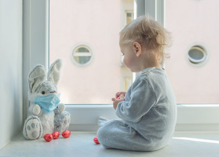 Easter 2020 concept with child in home quarantine playing at the window with his sick plush bunny wearing a medical mask against viruses during coronavirus COVID-2019 and flu outbreak.の写真素材