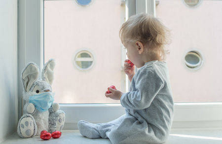 Easter 2020 concept with child in home quarantine playing at the window with his sick plush bunny wearing a medical mask against viruses during coronavirus COVID-2019 and flu outbreak.の写真素材