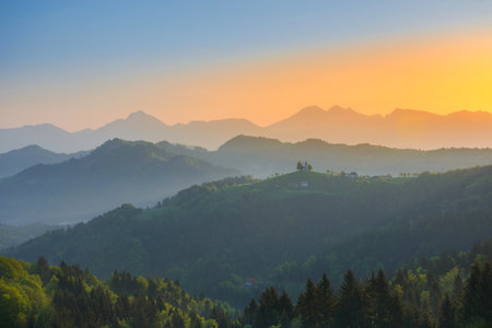 Slovenian breathtaking landscape at sunrise with Julian Alps and charming little church of Sveti Tomaz (Saint Thomas) on a hill, in spring. Beautiful misty morning in the mountains, in Slovenia.の写真素材