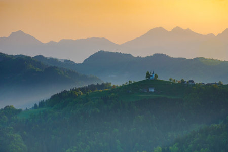 Slovenian breathtaking landscape at sunrise with Julian Alps and charming little church of Sveti Tomaz (Saint Thomas) on a hill, in spring. Beautiful misty morning in the mountains, in Slovenia.の写真素材