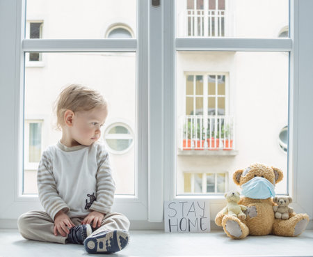 Child in home quarantine playing at the window with his sick teddy bear wearing a medical mask against viruses during coronavirus COVID-19 and flu outbreak, with the advice to stay home.の写真素材