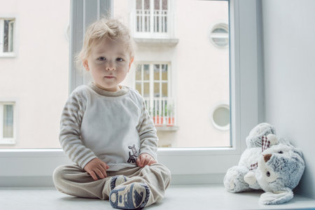 Cute boy at home looking through the window and playing with his toysの写真素材