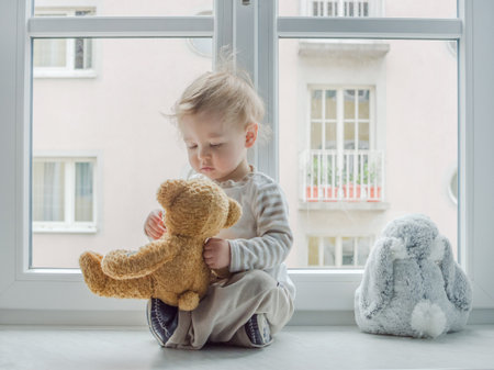 Cute boy at home looking through the window and playing with his toysの写真素材