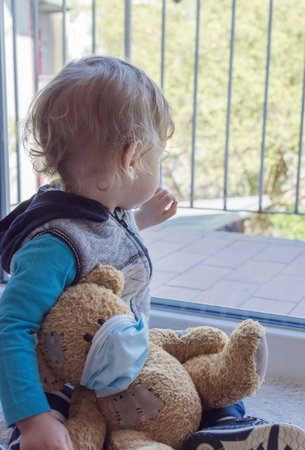 Child in home quarantine playing at the window with his sick teddy bear wearing a medical mask against viruses during coronavirus and flu outbreak. Children and illness COVID-2019 disease conceptの写真素材
