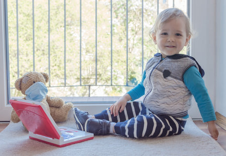 Child in home quarantine playing at the window with his sick teddy bear wearing a medical mask against viruses during coronavirus and flu outbreak. Children and illness COVID-2019 disease conceptの写真素材