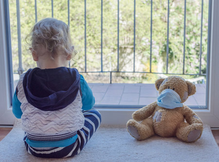 Child in home quarantine playing at the window with his sick teddy bear wearing a medical mask against viruses during coronavirus and flu outbreak. Children and illness COVID-2019 disease conceptの写真素材