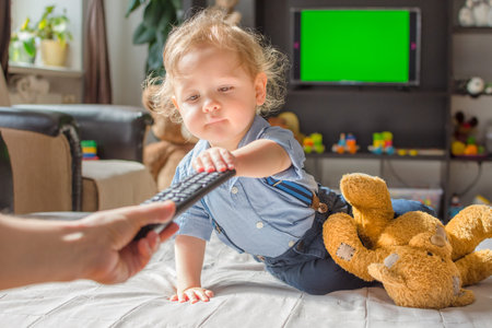 Cute baby boy taking the remote control to watch TV sitting on a couch in the living room at homeの写真素材